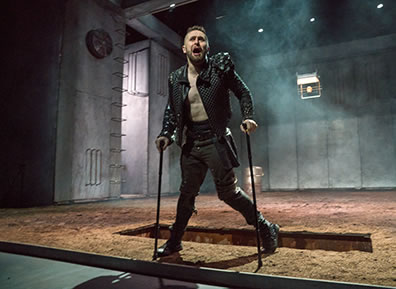 Production photo of Richard using his crutches as he stands in front of an open grave; his black Elizabethan jacket is unbuttoned showing his bare abdomen, and his left leg is crooked.